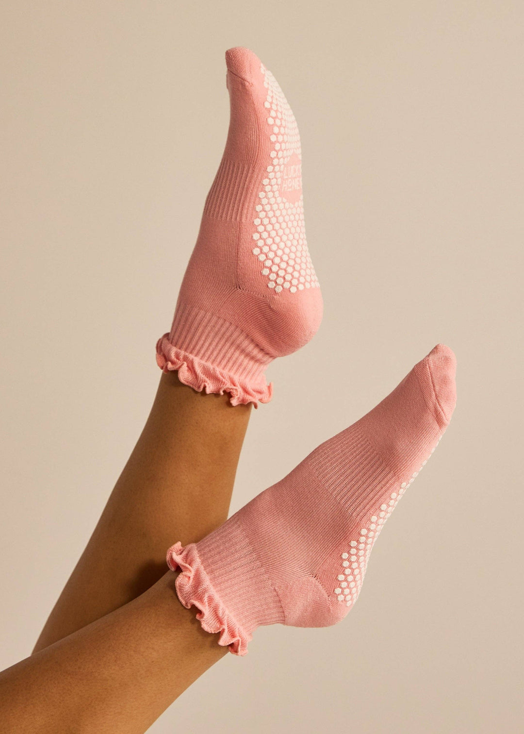A pair of pink ankle socks with white grip dots and ruffled cuffs, shown on a person's feet against a solid beige background.